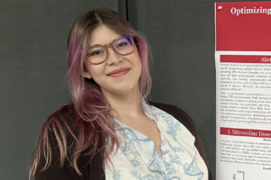 Anissa-landscape A person with shoulder-length pink-toned hair and glasses standing next to a scientific research poster.