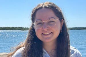 Kenna Berg A smiling woman with long brown hair and glasses stands in front of a body of water.