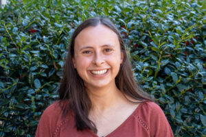 Melissa Rogers A smiling woman with long brown hair wearing a rust-colored top stands in front of a green hedge.